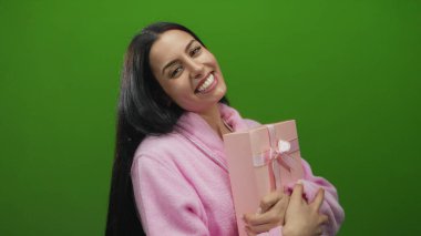 Parlak yeşil bir arka planda pembe hediye kutusunu tutan ve kutlayan kadın, rahat pembe bir bornozla mutluluk ve neşe ifade ediyor, neşeli ve neşeli bir atmosfer yaratıyor..
