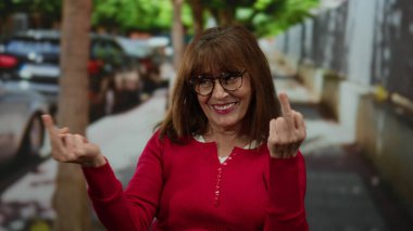 Kırmızı kazaklı İspanyol bir kadın şehir arka planında orta parmaklarıyla bir jest yapıyor, duygularını ve davranışlarını açık havada ifade ediyor..