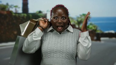 Kumsal girişine şaşıran bir kadın, güneşli bir günde elinde okyanus ve palmiye ağaçlarıyla birlikte açık havada alışveriş torbaları tutuyor..