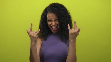 Kıvırcık saçlı ve kollarını kaldırmış genç bir kadın sarı stüdyo ortamında rock boynuzları gösteriyor..