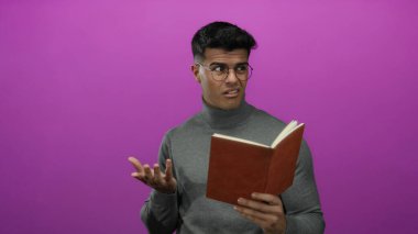 Gözlük takan ve gri kazak giyen genç adam pembe arka plana karşı kitap okuyor. Stüdyo ortamında düşünceli ve meraklı olduğunu ifade ediyor..