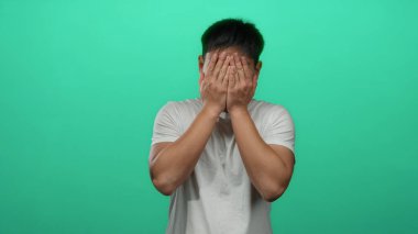 İzole edilmiş yeşil arka plan duvarındaki genç Çinli adam, elleri ile gözleri örtüp açarken neşeli, neşeli bir anı videoda yakalıyor..