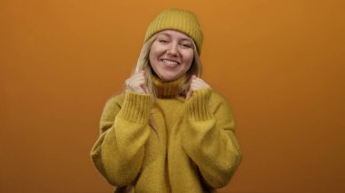 Sıcak sarı bir kazak ve bere içinde neşeli bir şekilde gülümseyen genç sarışın kadın, canlı turuncu bir arka planda izole edilmiş, olumlu duygular ve şık kış tarzı sergiliyor..
