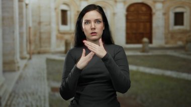 Esmer kadın üniversite kampüsünde etkileyici bir şekilde el kol hareketi yapıyor. Geçmişinde tarihi bir mimari var.