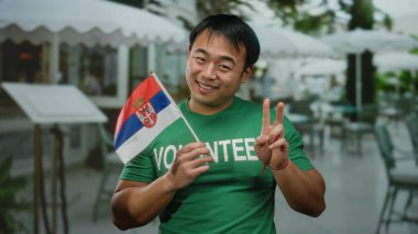 Terasta Sırp bayrağı taşıyan genç bir adam yeşil gönüllü tişörtü giyerken şehir ortamında neşeli bir ifade ve barış hareketi sergiliyor..