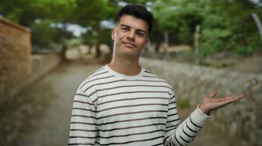 Çizgili tişörtlü ve elini açık bir parkta dikilen genç bir adam. Arka planda taştan bir yol ve yemyeşil bir zemin var. Neşeli görünüyor..