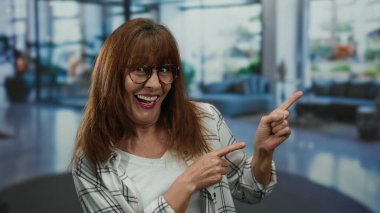 Üst düzey İspanyol kadın gülümseyerek ve kapalı bir otel lobisinde gözlük ve ekose gömlek takarak neşeli ve davetkar bir atmosfer sunuyor..