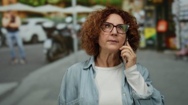 Şehir caddesinde akıllı telefondan konuşan bir kadın gözlük takıyor ve açık havada günlük giysiler giyiyor. Modern yaşam tarzını ve orta yaş iletişimini kaydediyor..
