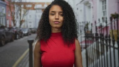 Kırmızı bluzlu ve kıvırcık saçlı genç bir kadın demir çitlerle ve park edilmiş arabalarla dolu bir sokakta kendini işaret ediyor..