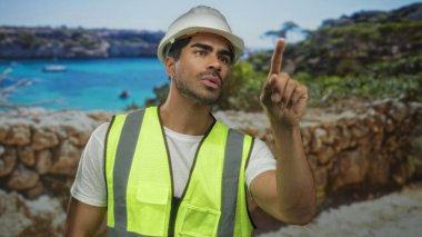 Beyaz kasketli ve neon yelekli adam teftiş sırasında kayalık deniz duvarının yakınındaki caddeyi işaret ediyor..