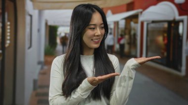 Sokak ortamında gülümseyen bir kadın, açık ellerle sunuyor, günlük kıyafetler giyiyor, bulanık dükkanlarla çevrili, mutluluk ve şehir hayatını aktarıyor..