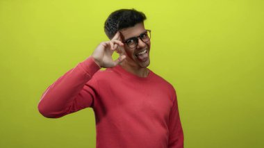 Gözlüklü genç adam pembe kazak giyiyor. Parlak sarı arka plana karşı neşeli poz veriyor. Kendine güveni, mutluluğu ve modern tarzı gösteriyor.