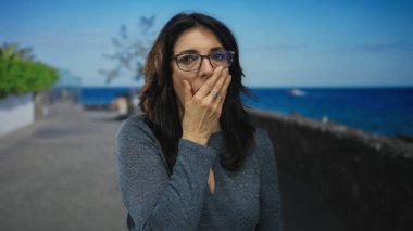 Gözlüklü orta yaşlı bir kadın güneşli deniz kenarında gezinti caddesinde el kol hareketi yapıyor; sessizlik..