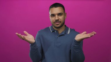Mavi polo tişörtlü bir adam stüdyoda, sanki seçenekleri tartarmış gibi şaşkın bir ifadeyle avuç içlerini havada tutuyor..