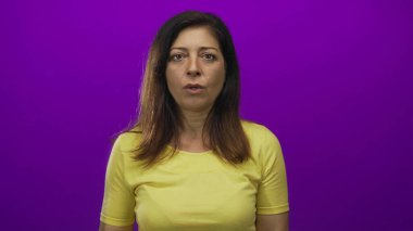 Latin kökenli orta yaşlı bir kadın mor bir stüdyoda eli ile ağzını kapatıyor sarı gömlek ve geniş gözler giyiyor..