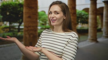 Genç beyaz kadın açık avuçlar ve canlı yeşil parktaki taş sütunlar tarafından samimi bir gülümseme sergiliyor; neşe.