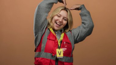 Kırmızı yelekli, gülümseyen gönüllü kadın elleri kahverengi arka planda kalp şekli yapıyor..