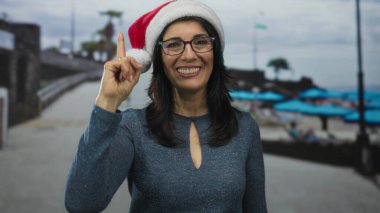 Noel Baba şapkası takan ve gözlük takan kadın deniz kenarındaki şemsiyelerin olduğu plaj yolunda parmağını yukarı kaldırıyor; bayram neşesi.