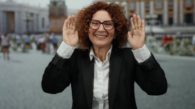 Orta yaşlı, siyah takım elbiseli, gülümseyen İspanyol kadın arka planda hareketli şehir hayatı olan Aziz Peter Meydanı, Vatikan 'da duruyor..