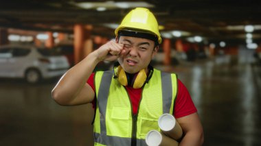 Yüksek görüş açısına sahip yeleği ve kaskı olan genç bir adam elinde bina içi park alanının planlarıyla ağlarken şaka yapıyor..