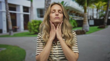 Güneşli bir günde dışarıda dikilen bir kadın boğazını tutuyor ve modern bir sokak ve yeşillik geçmişiyle pek iyi görünmüyor. Potansiyel sağlık sorunları olduğunu gösteriyor..