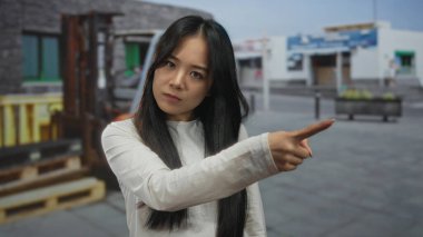 Beyaz tişörtlü genç Çinli kadın sokak sahnesinde, modern şehir arkaplanıyla saygısız bir jest yapıyor. Kararlılık ve güven gösteriyor..