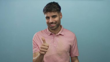 Latin kökenli genç adam gülümser ve açık mavi bir stüdyo ortamında başparmak kaldırır; güven pozitifliği başarı teşviki.