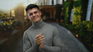 Genç İspanyol adam gülerek ve alkışlayarak bir şehir caddesinde, gözlük ve balıkçı yaka giyerek, şehir dışında bir yerde neşe ve sıcaklık anı yakalıyor.