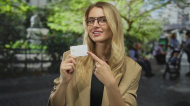 Parkta elinde kredi kartıyla gezen yeşil ağaçlı gözlük takan bir kadın. Arka planda bulanık duran insanlar, güneşli bir günü gösteriyor..