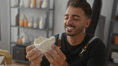 Adam stüdyoda, el yapımı gülümseyen seramik damarların arasında iki eliyle kil kase tutuyor..