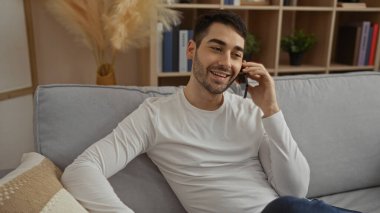 Genç İspanyol bir adam, evin içinde bir kanepede dinleniyor, telefonda gülümsüyor, ev dekorasyonu ve modern bir apartman dairesinde kitap raflarıyla çevrili..