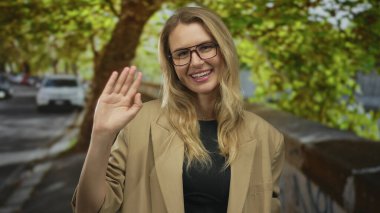 Gülümseyen ve el sallayan bir kadın gözlüklü ve bej ceketli bir kadın. Benekli güneş ışığı altında, açık havada sıcak ve rahatlatıcı bir gün olduğunu gösteriyor..