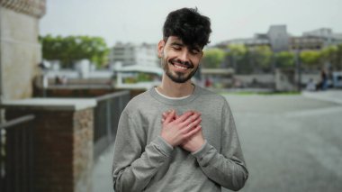 Sokak ortamında göğsünde elleri göğsünde sıcacık gülümseyen genç bir adam..