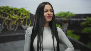 Uzun saçlı İspanyol bir kadın yeşil bitkilerle dolu bir parkta reddedilme jesti yapıyor ve arka planda duyguları ve açık hava ortamını gösteren taş bir yol var..