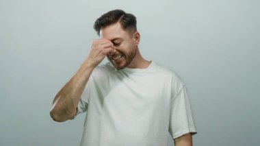 Beyaz gömlekli sakallı genç adam, beyaz arka planda dikilirken burun köprüsünü çimdiklerken hayal kırıklığı ve tefekkür ifadesini gösteriyor..