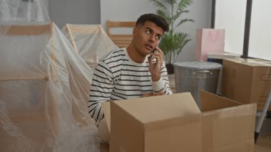 Çizgili gömlekli genç adam, yeni bir apartman dairesinde karton kutuları açarken telefonla konuşuyor. Yeni bir taşınma ya da taşınma işlemi öneriyor..