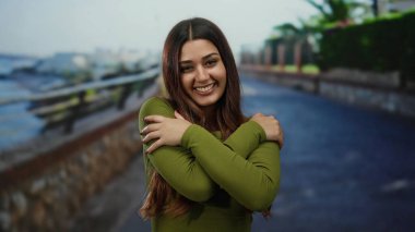 Güneşli bir sahil yürüyüşünde gülümseyen bir kadın okyanusun arka planında neşe ve dinginlik sergiliyor.