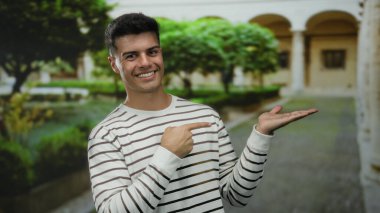 Üniversite bahçesinde jest yapan çizgili tişörtlü genç bir adam akademik ortamda olumlu ifade sergiliyor.