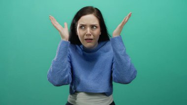 Şaşkınlık ve şaşkınlık ifade eden bir kadın canlı yeşil bir arka plana karşı, mavi bir kazak giyen, esmer oyuncu ve meraklı duyguları vurgulayan, geniş gözlü, eller kaldırılmış.