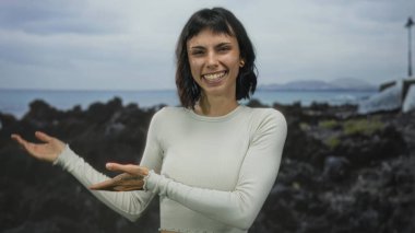 Beyaz ekin giyen kadın, kayalık sahil kenarında uzanmış kolları ve avuç içleri ile yumuşak jestler; davet yansıma dinginliği açıklığı.