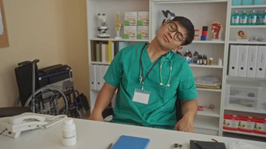 Tıbbi önlüklü genç adam bir klinikte oturuyor, stresli görünüyor, boynunda steteskop ve arka planda tekerlekli sandalye var..