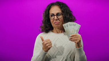 Kafası karışık bir ifadeyle orta yaşlı bir kadın, mor bir arka planda, baş parmağıyla kenarı işaret ederken, bir yandan kirli para tutuyor..