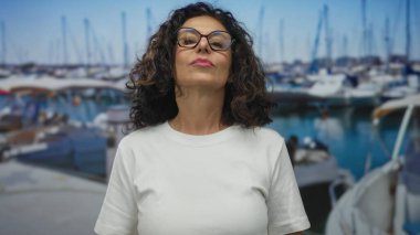 Deniz kıyısında gözlüklü bir kadın, elini kalbinin üzerine koyuyor, bir tekne arkaplanının ortasında, açık hava ortamında düşünceli bir yüz ifadesi öneriyor..