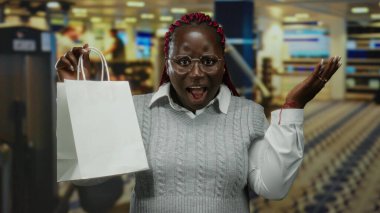 Gözlüklü ve süveterli bir kadın alışveriş torbası tutuyor. Arka planda bulanıklık var. Modern bir mağaza ya da alışveriş merkezi ortamı öneriyor..
