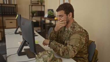 Odanın içindeki dağınık ofis masasında ağzını kapatan genç İspanyol asker monitörler ve dosyalarla dolu..