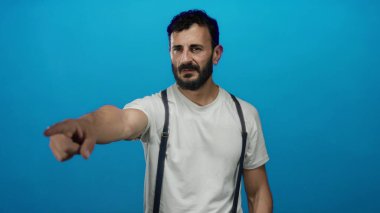Mavi arka planda sakallı İspanyol bir adam beyaz gömlek ve pantolon askısı giyiyor, odak ve yönünü modern bir ortamda aktarıyor..