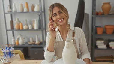 Kadın çanak çömlek atölyesinde bitmemiş vazo ile dönen kil çarkın yanında telefonu kulağına tutuyor; iletişim yaratıcılığı.