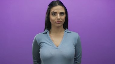 Mor bir yüzle mor bir duvara karşı duran, mavi gömlekli, doğrudan kameraya bakan, genç ve kendinden emin, kapalı, dışa dönük, anlamlı, canlı renklerde bir kadın..