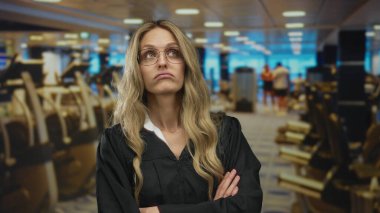 Spor salonunda kollarını kavuşturmuş bir kadın sinirli görünürken, üniforma giyen genç sarışın yargıç içeride spor salonu duvarının yanında duruyor..