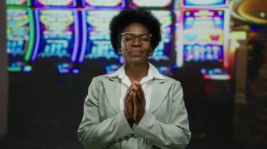 Kumarhanede elinde kumar makineleri, gözlüklü ve takım elbiseli bir kadın, elleri birarada, odak ve iç mekan renklerine karşı düşünme tasviri yapıyor..